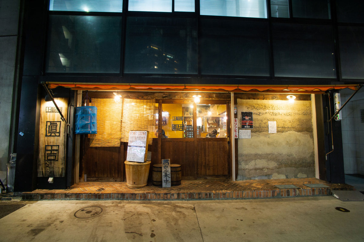 魚まみれ眞吉 原宿店の夜の店先。左に「眞吉」と書かれた木の看板、オレンジ色の庇、すだれ、樽の上に「営業中」の札、ガラス戸の奥に灯りのついた店内。