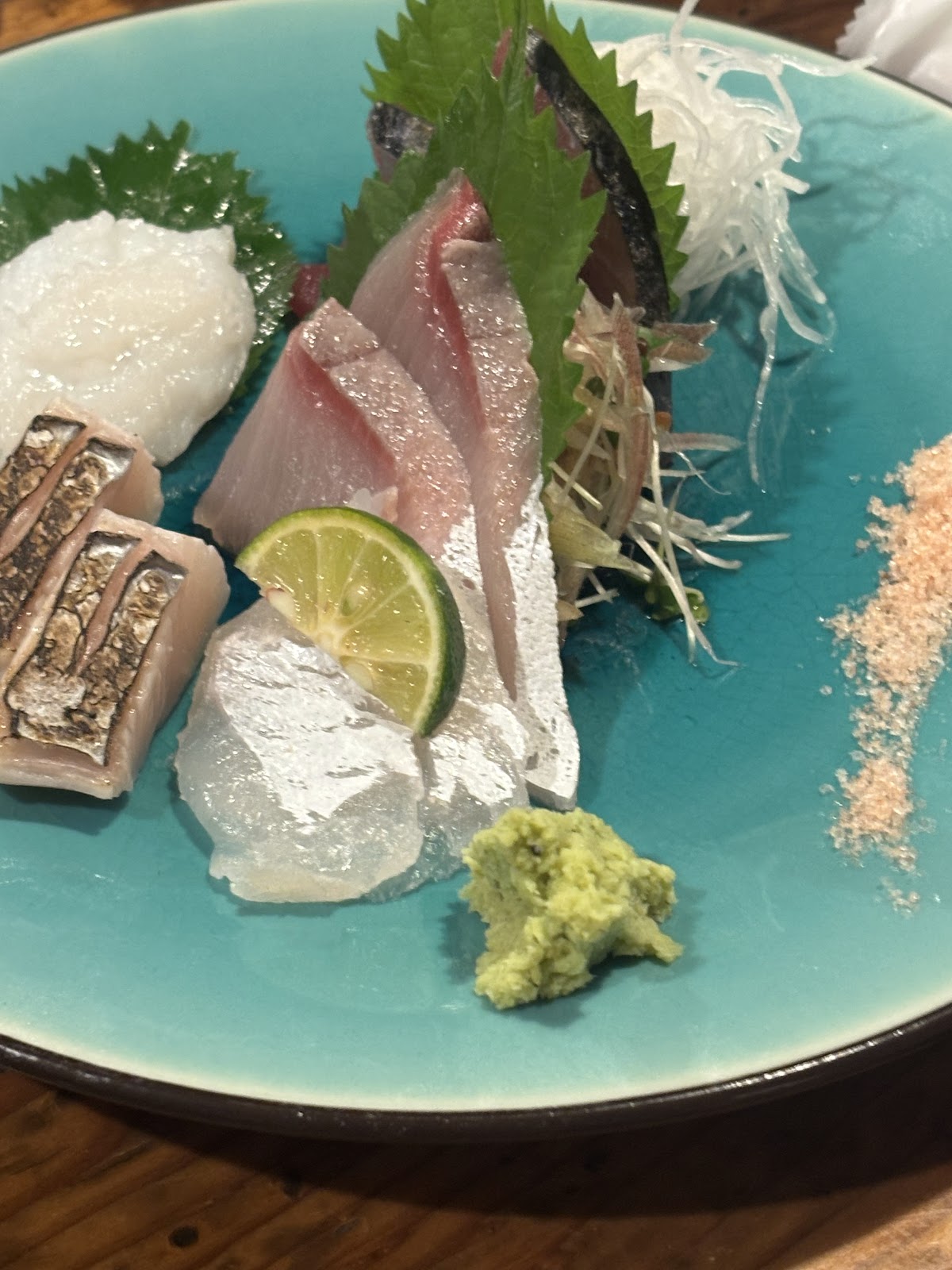 青いガラスの皿に盛った刺身。皮目を炙った魚、白身、脂ののった切り身、大葉、大根のつま、すだちの輪切り、わさびと桃色の塩。