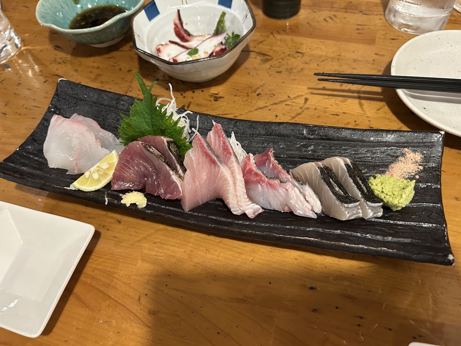 木のカウンターに置いた黒い長皿の刺身盛り。白身、赤身、炙り、大葉とつま、すだち、わさび。奥にたこの小鉢と箸。